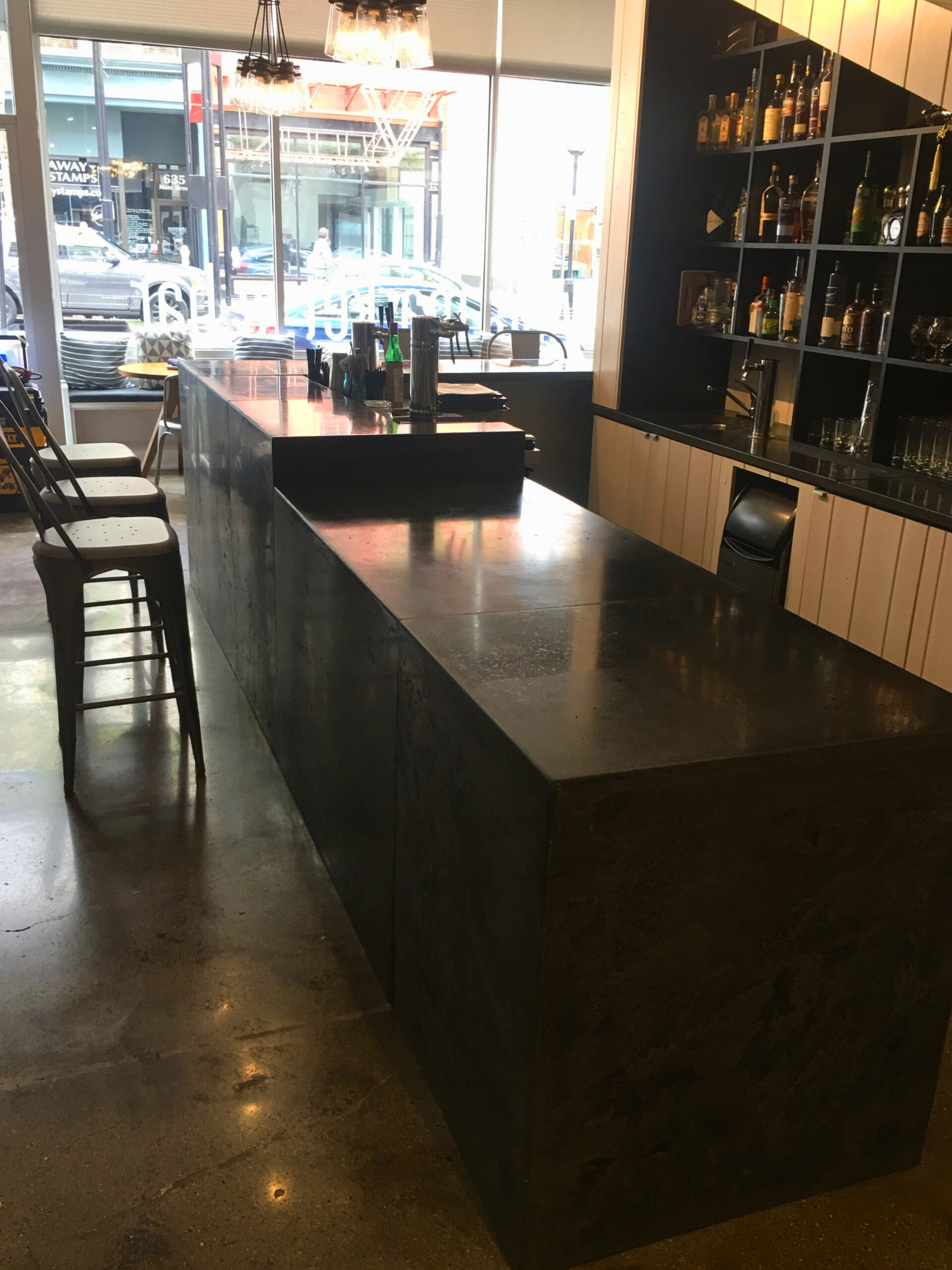 Modern bar interior with dark counter seating, black bar stools, and shelving stocked with bottles, Cincinnati establishment with street-facing windows