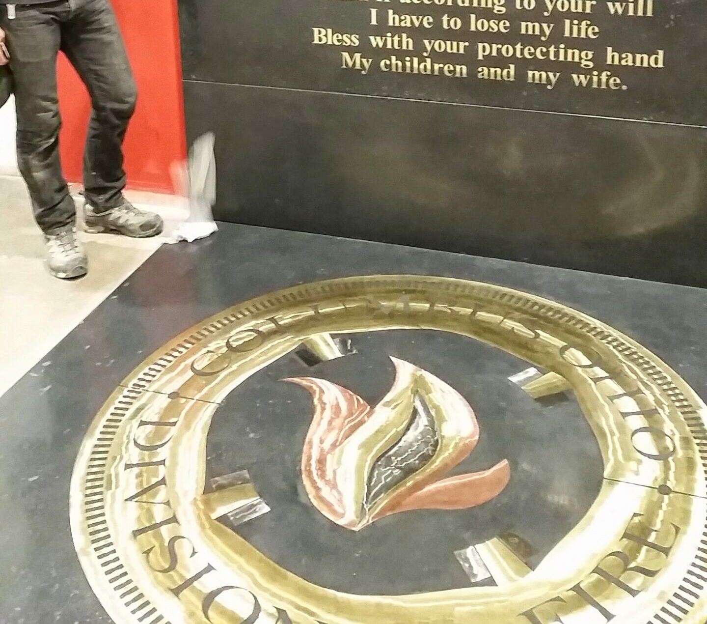 Person standing beside firefighter's prayer plaque and brass seal with flame emblem at Cincinnati firehouse